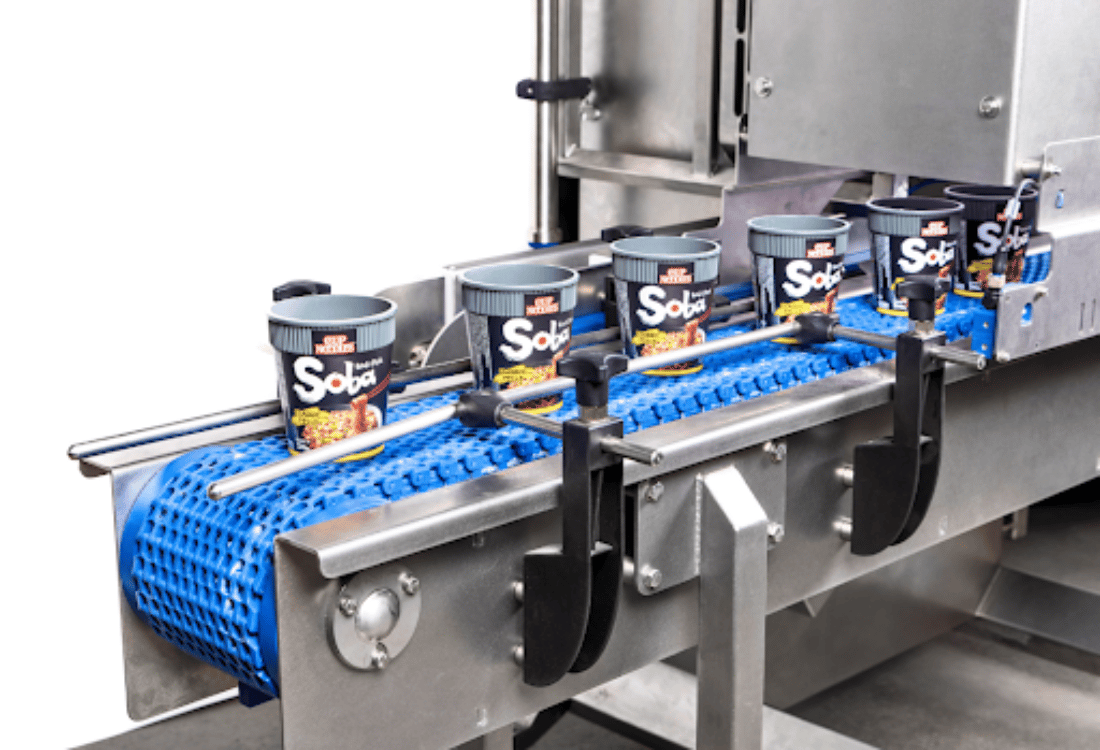 A line of ready-to-eat noodle cups moves along a blue conveyor belt, guided by automated machinery.